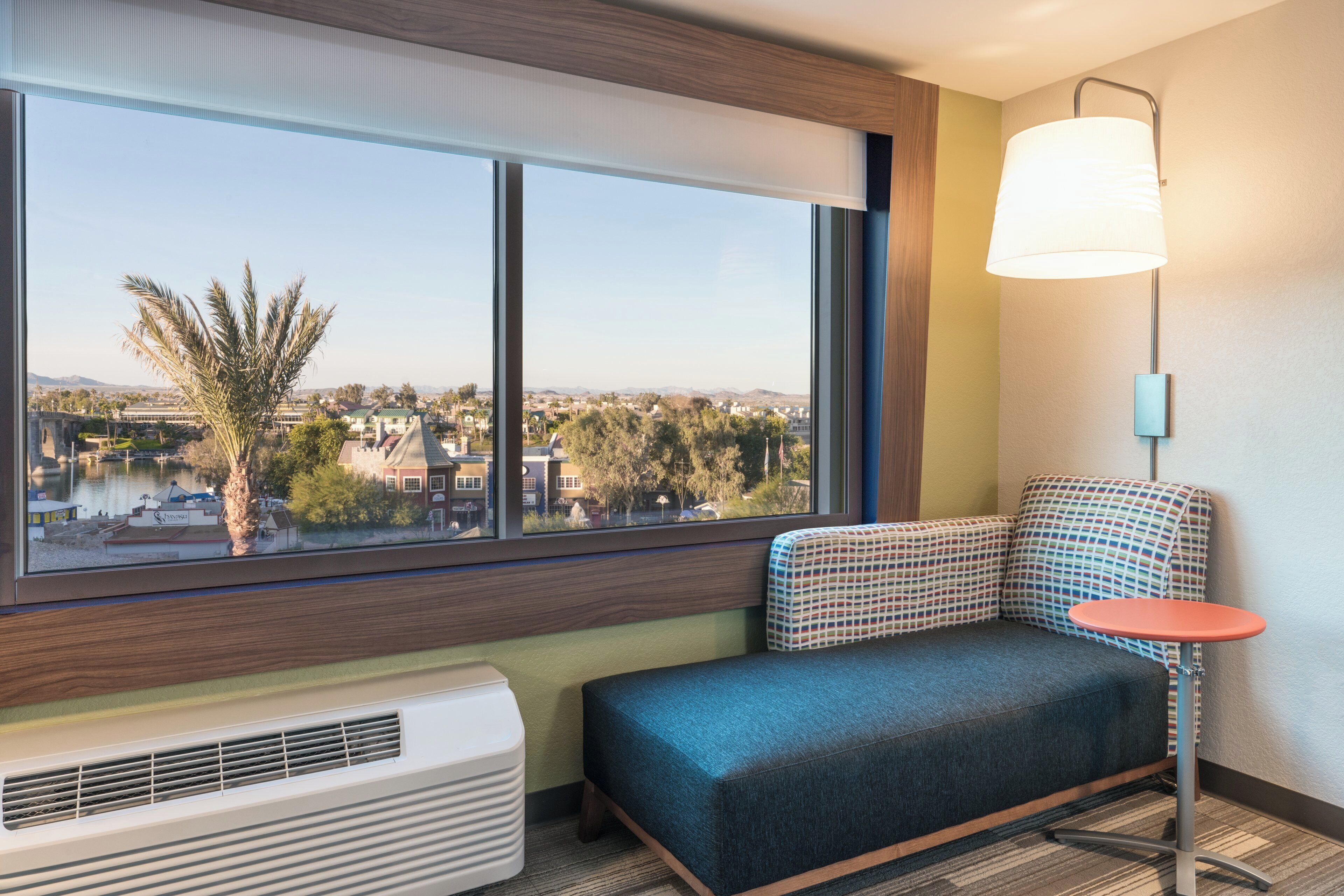 A hotel room with a large window, a sofa, a small table, and a wall-mounted lamp.