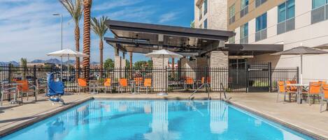 A swimming pool with lounge chairs and umbrellas, surrounded by a fence and palm trees.
