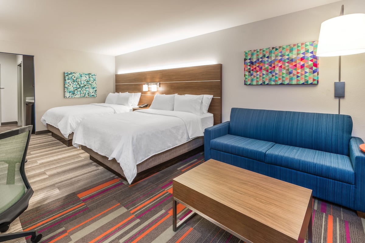 Une chambre d’hôtel avec un grand lit, un canapé bleu, une table basse en bois et un tableau abstrait aux couleurs vives accroché au mur.