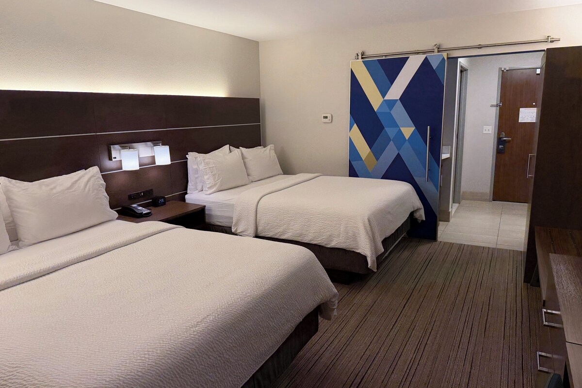 A hotel room with two beds, a nightstand with a phone, a wall-mounted light, and a door with a geometric-patterned curtain.
