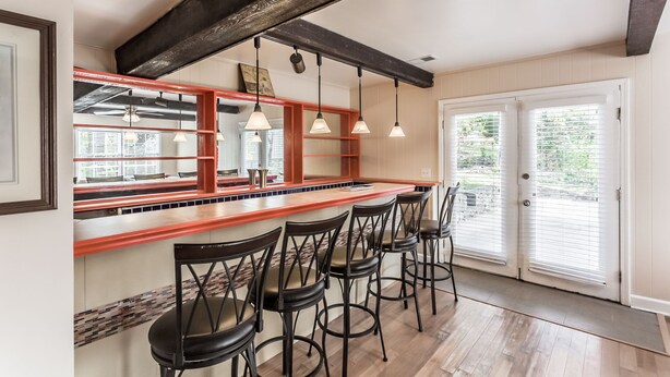 Una cocina con barra de madera, sillas negras, lámparas colgantes y vistas por la puerta.