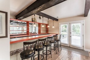 A kitchen with a wooden bar, black chairs, pendant lights, and a view through the door.