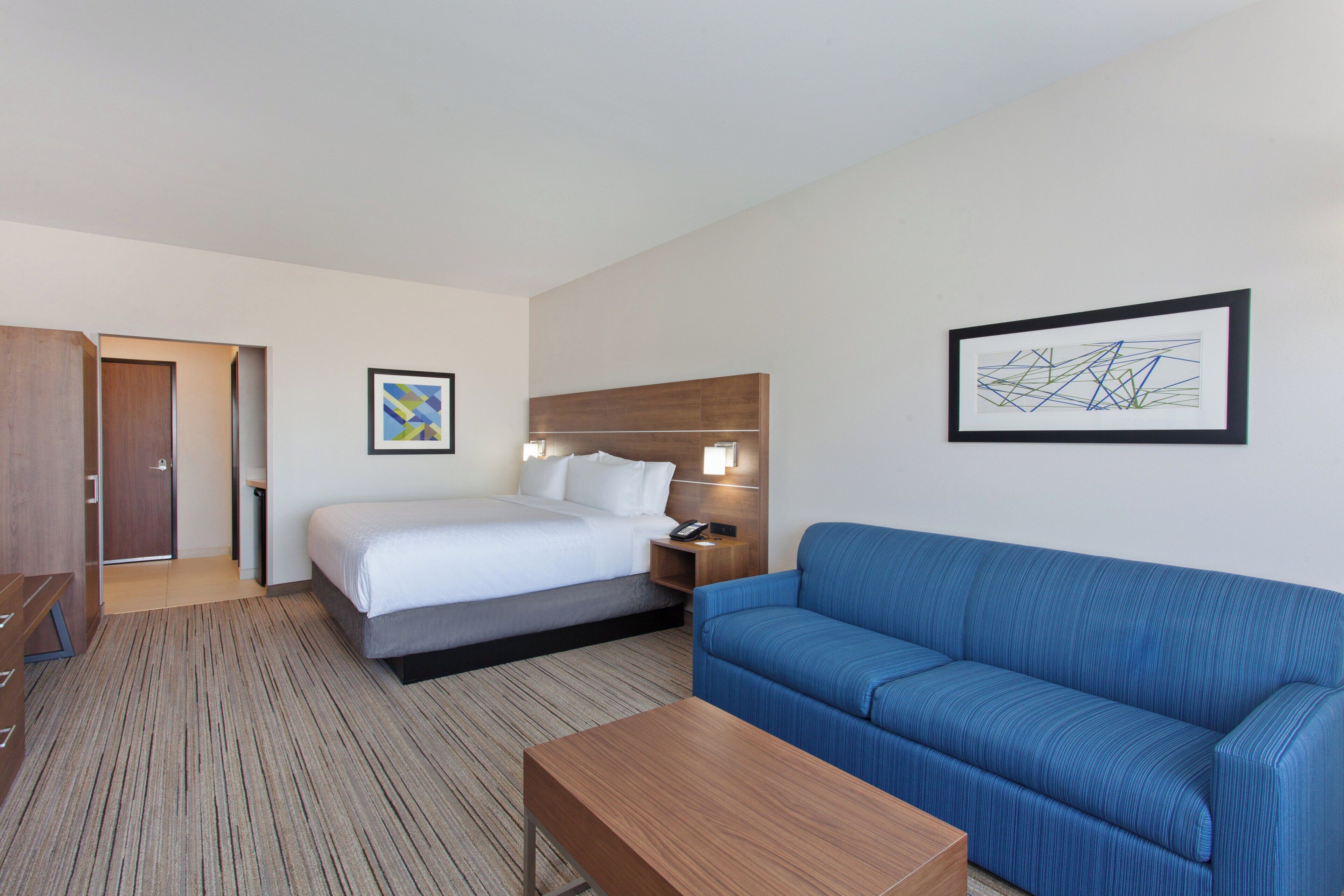 A hotel room with a bed, a blue sofa, a wooden coffee table, a nightstand, a lamp, and two framed artworks on the wall.