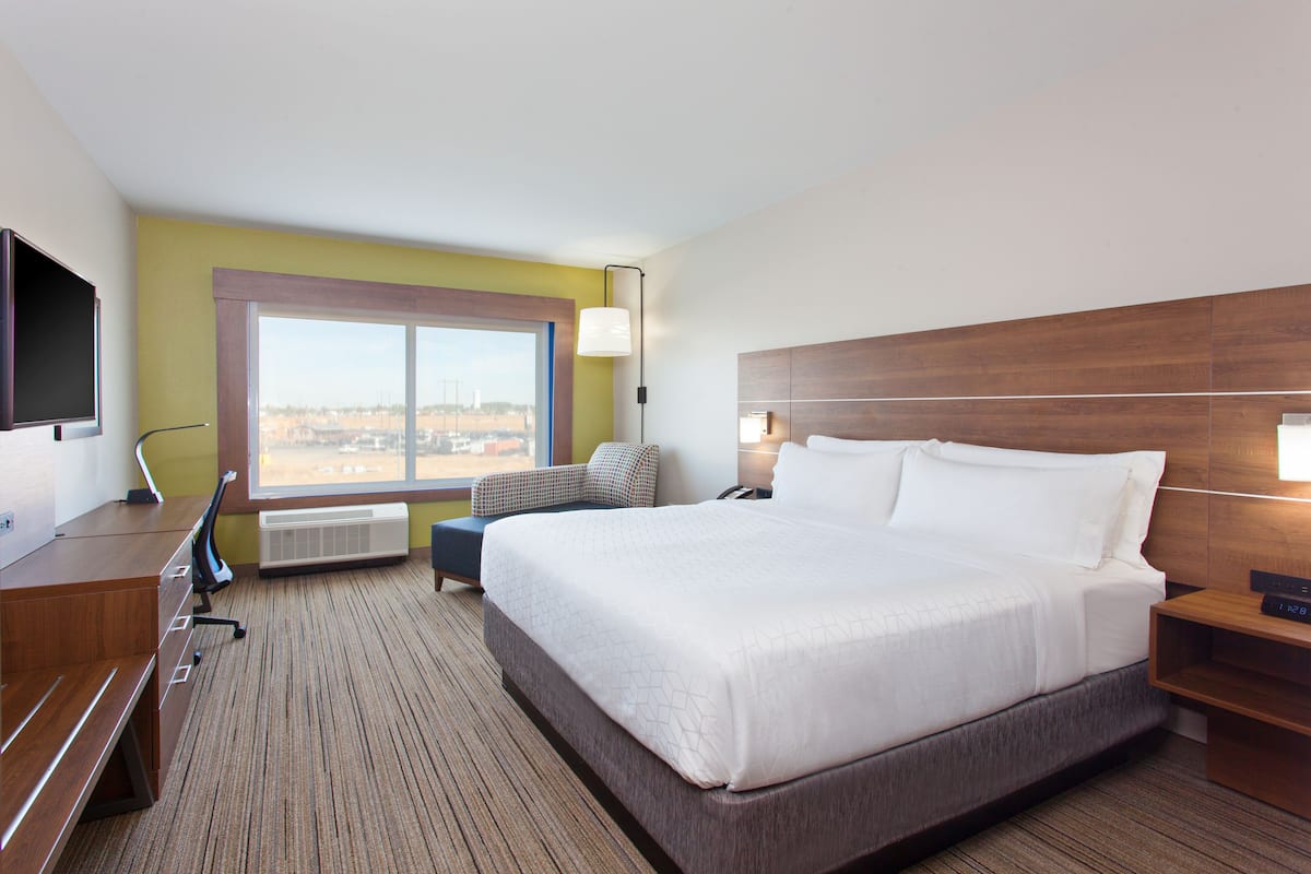 A hotel room with a large bed, a desk with a chair, a flat-screen TV, a window with a city view, and a wall-mounted lamp.