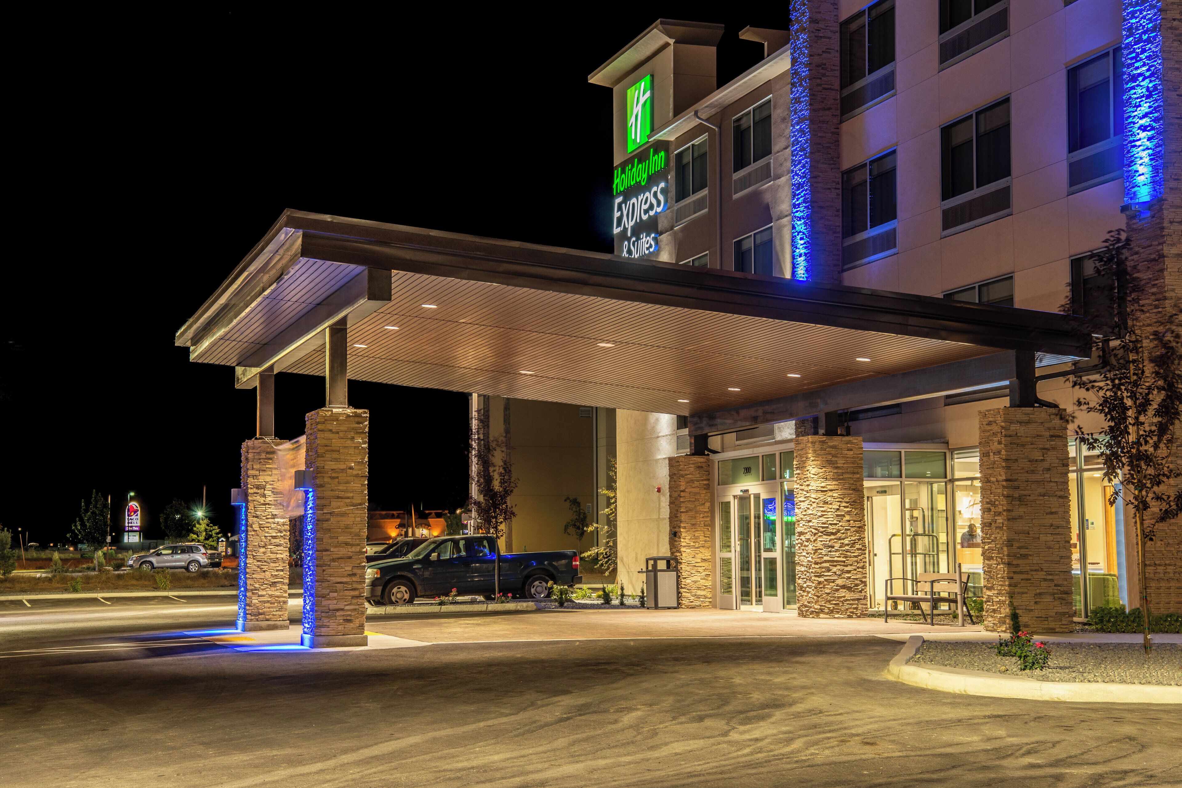 Een Holiday Inn Express & Suites gebouw met een overdekte ingang, stenen pilaren en een blauw verlichte gevel.