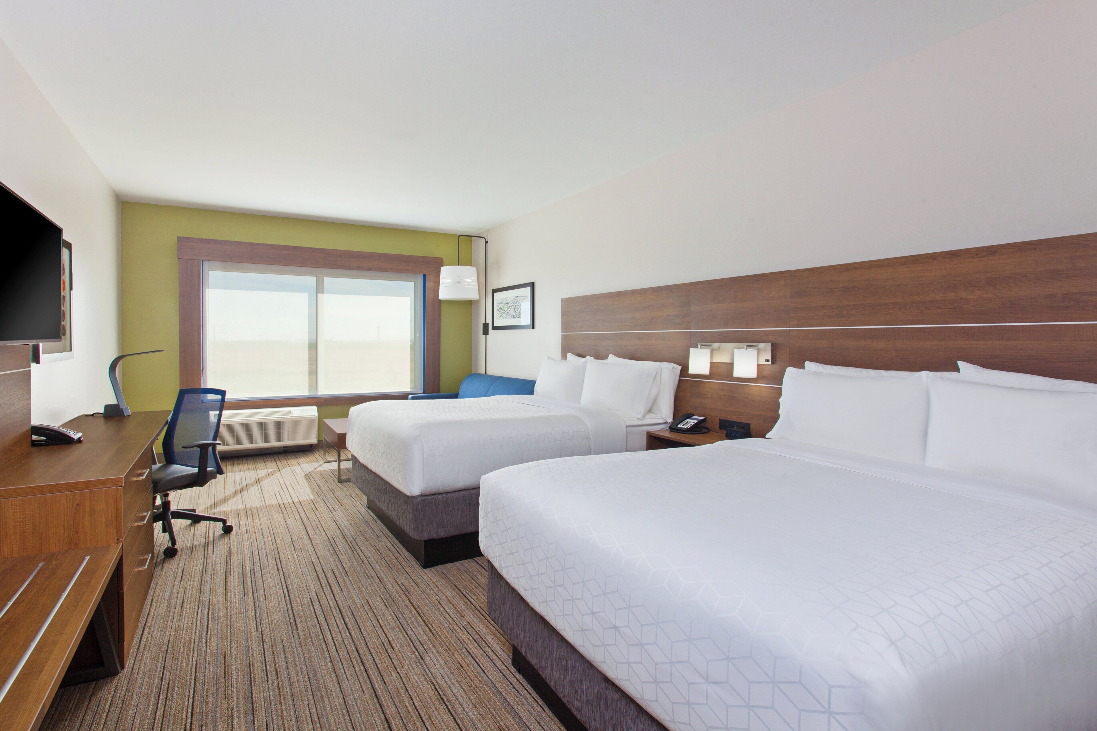 A hotel room with two beds, a desk with a chair, a flat-screen TV, a bedside table with a lamp, and a window with a view.