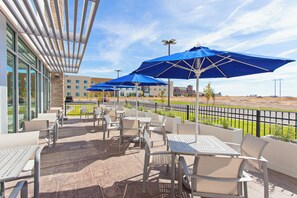 Un espace extérieur aménagé en terrasse, avec des tables et des chaises, des parasols bleus et une vue sur un bâtiment en arrière-plan.