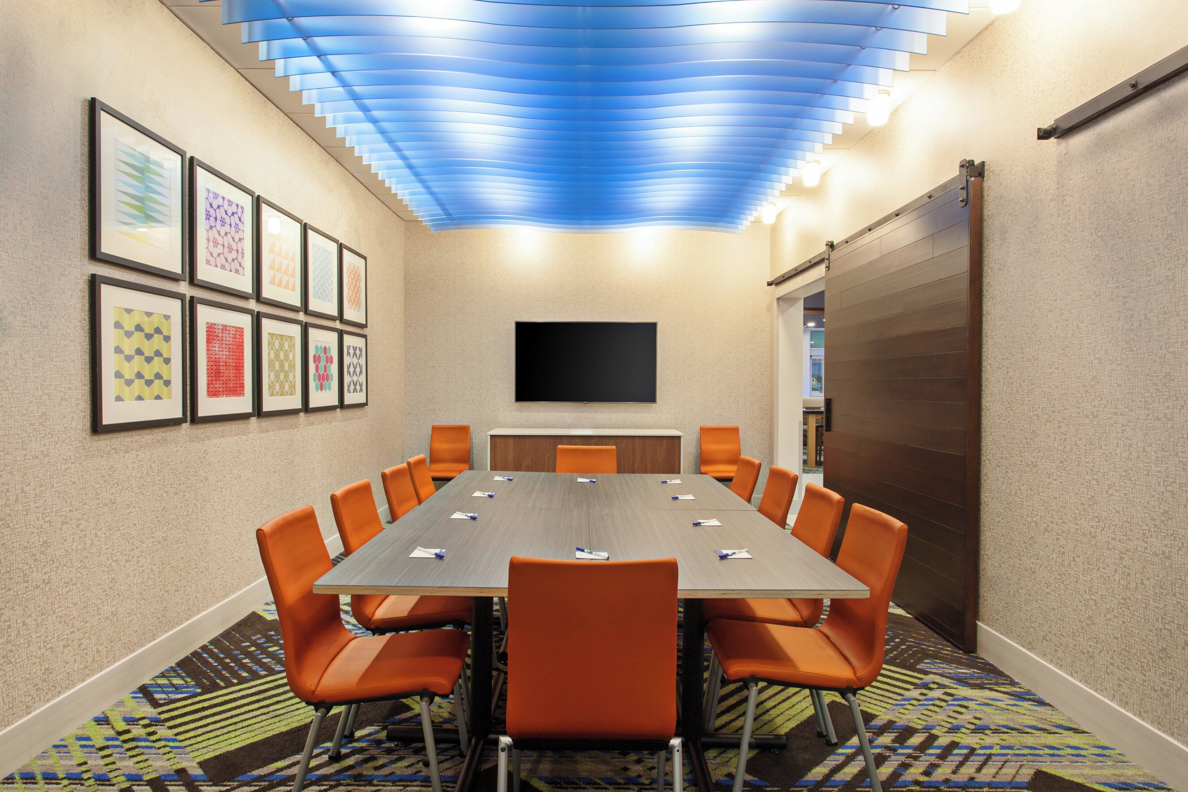 A conference room with a long table, orange chairs, framed artwork on the wall, and a flat-screen TV mounted above the table.