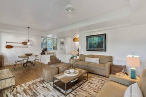 A spacious living room with a sofa, coffee table, and dining area. There is a staircase leading to an upper level, and a painting on the wall.