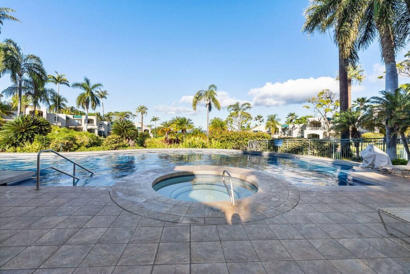 A swimming pool with a hot tub in the center, surrounded by palm trees and a tiled deck.