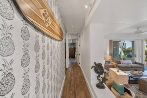 Ein Flur mit Tapete mit Ananasmuster, einer Holzbalkendecke und Blick in einen Wohnbereich mit Sofa und Schildkrötenskulptur.