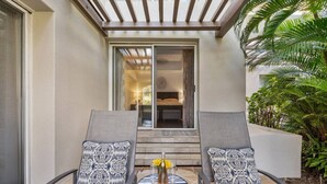 Un espace patio avec deux fauteuils de jardin gris, une petite table ronde ornée d’un vase et d’un verre, et une vue sur une chambre à coucher à travers une porte vitrée.