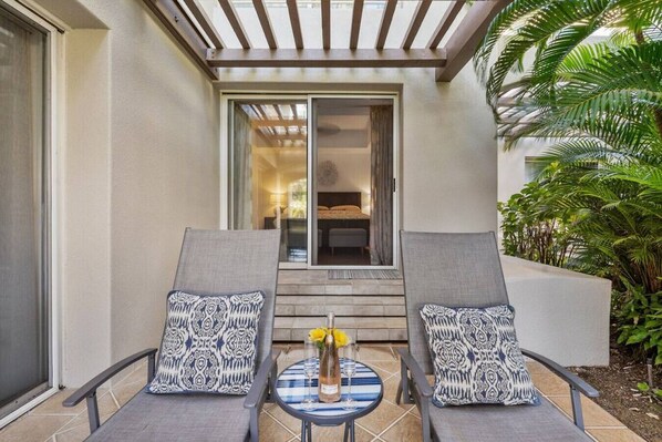 Une terrasse avec deux fauteuils de jardin gris, une petite table sur laquelle est posé un vase rempli de fleurs jaunes, et une vue sur une chambre à coucher à travers une porte vitrée.