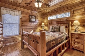 A wooden cabin bedroom with a bed, two bedside tables, a nightstand, a ceiling fan, and a window with blinds.