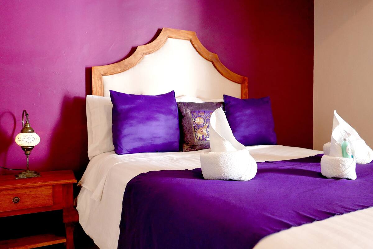 A neatly made bed with purple bedding, a wooden headboard, and a nightstand with a lamp.