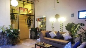 Una sala de estar con sofá, mesa de centro y plantas en macetas. Se observa una estantería y una lámpara fijada a la pared.