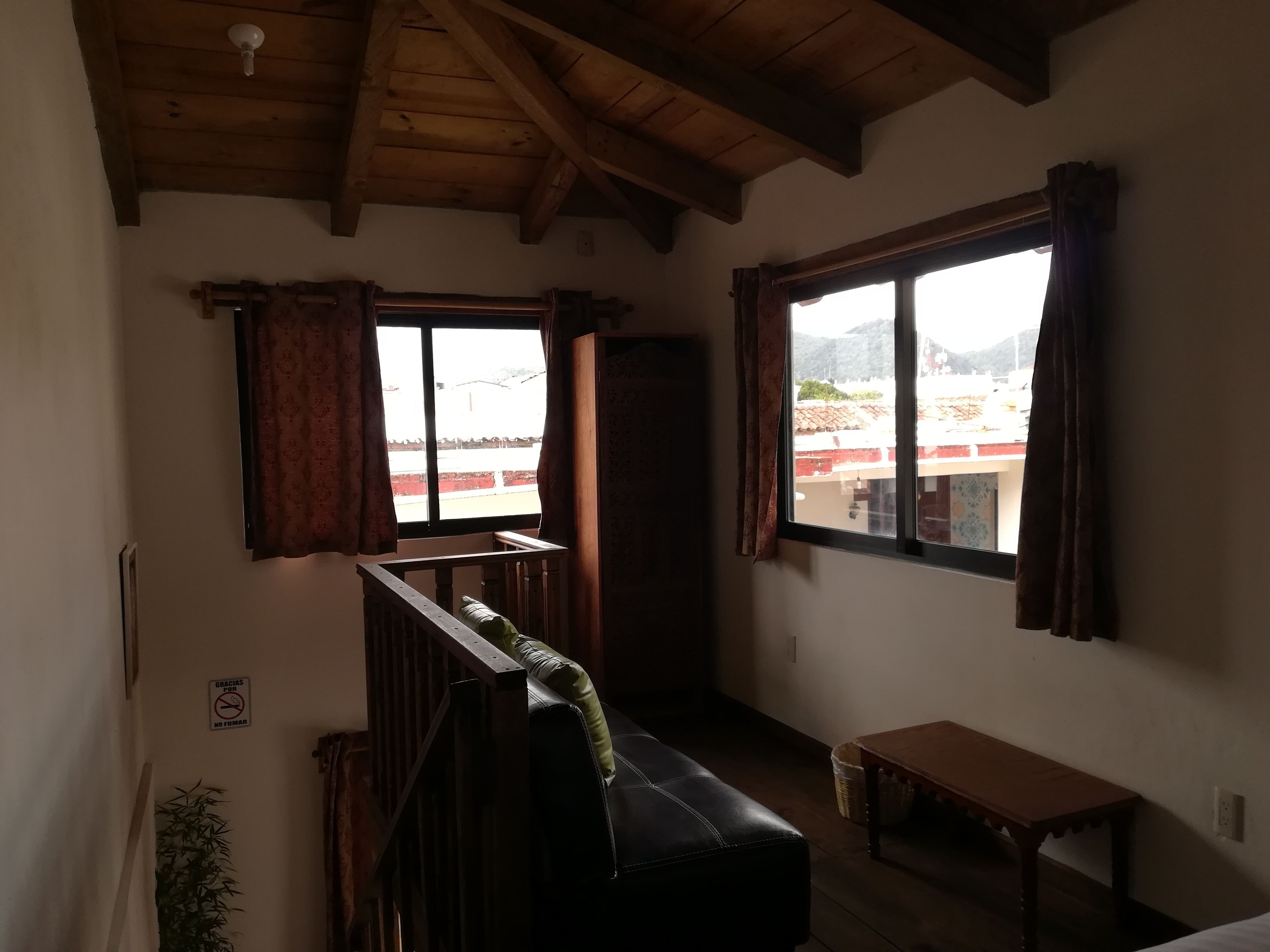 A room with a wooden ceiling, a black leather bench, a small wooden table, and two windows with curtains.