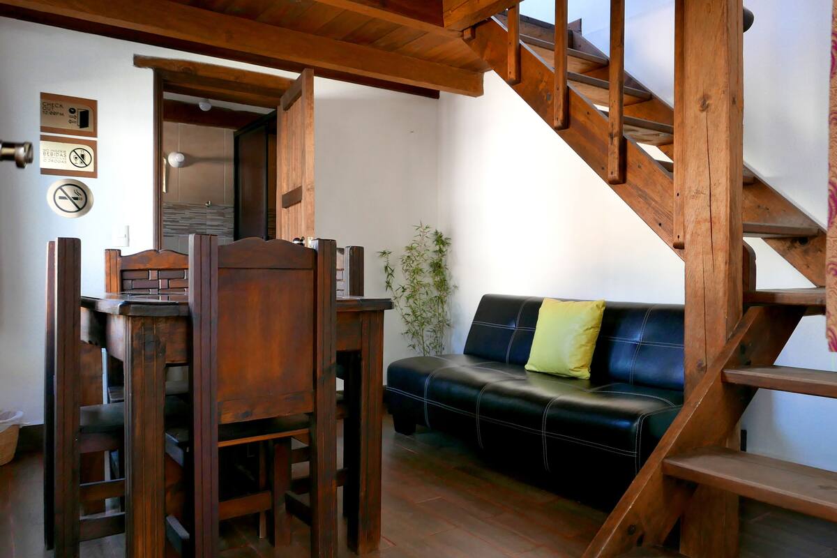 A wooden staircase leading to a loft area with a black leather sofa and a wooden dining table with chairs.