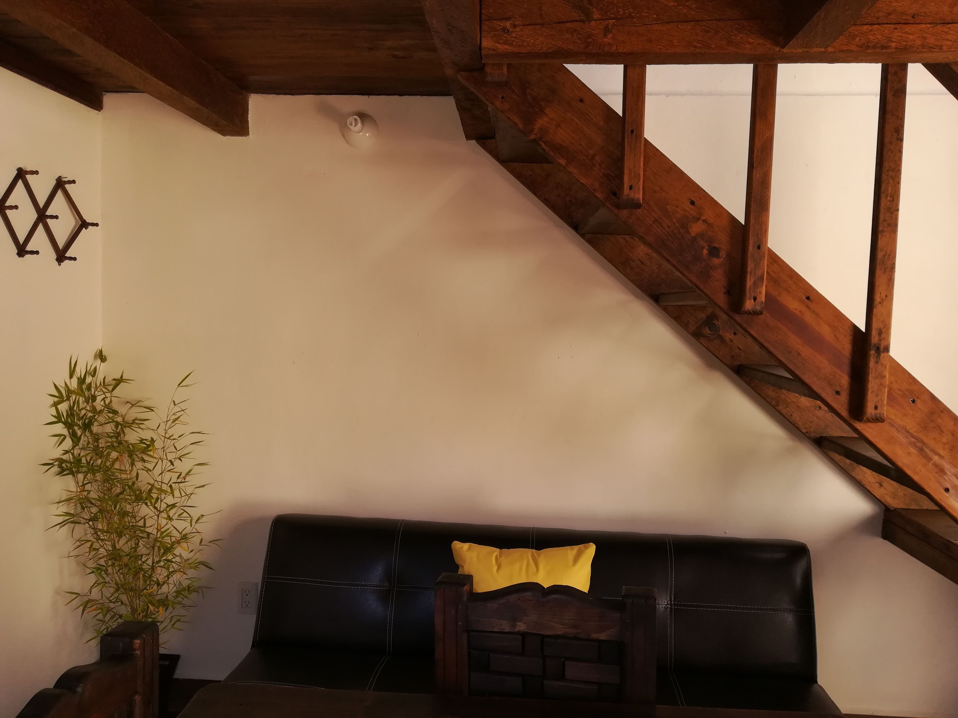 A room with a wooden staircase, a black leather sofa with a yellow pillow, and a bamboo plant.