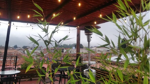 Un espacio de esparcimiento al aire libre con plantas de bambú, mesas y sillas, bajo una pérgola de madera con luces colgantes.
