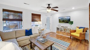 A living room with a sofa, coffee table, and a wicker chair. There is a ceiling fan, a TV mounted on the wall, and a window with blinds.