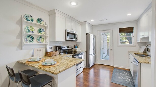 A modern kitchen with white cabinets, a granite countertop, a stainless steel refrigerator, and a microwave. There are two bar stools and a chalkboard sign on the countertop.