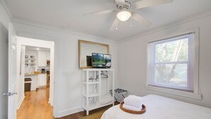 A bedroom with a bed, a ceiling fan, a TV on a stand, a window with blinds, and a view of a kitchen and bathroom through an open door.