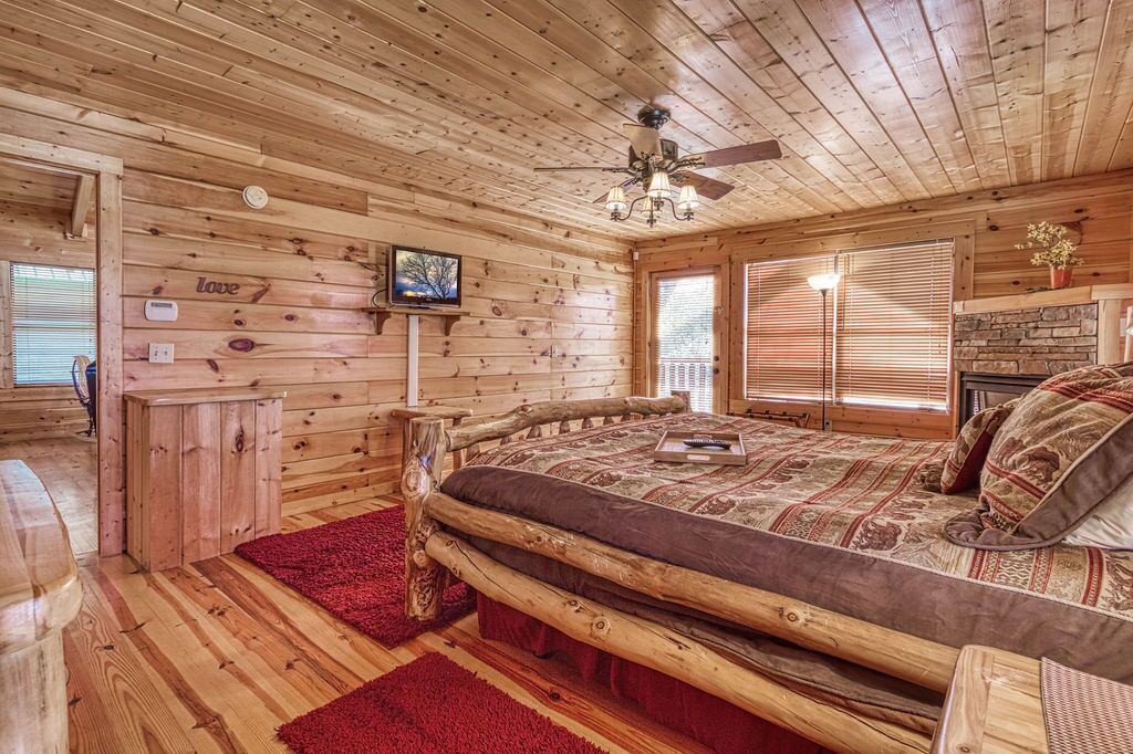 A wooden cabin bedroom with a bed, a ceiling fan, a fireplace, and a red rug.