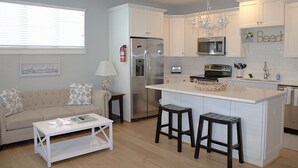 A modern kitchen with white cabinets, a stainless steel refrigerator, a microwave, and a stove. There is a small island with stools, a white coffee table, and a sofa with patterned cushions. A fire extinguisher is mounted on the wall. - 2BR Virginia Beach townhouse with king suite, balcony, WiFi, parking. (Virginia Beach)