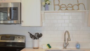 A kitchen with white cabinetry, a stainless steel microwave, a built-in oven, a sink with a modern faucet, and a shelf with a vase of flowers and the word "Beach" spelled out in rope. - 2BR Virginia Beach townhouse with king suite, balcony, WiFi, parking. (Virginia Beach)