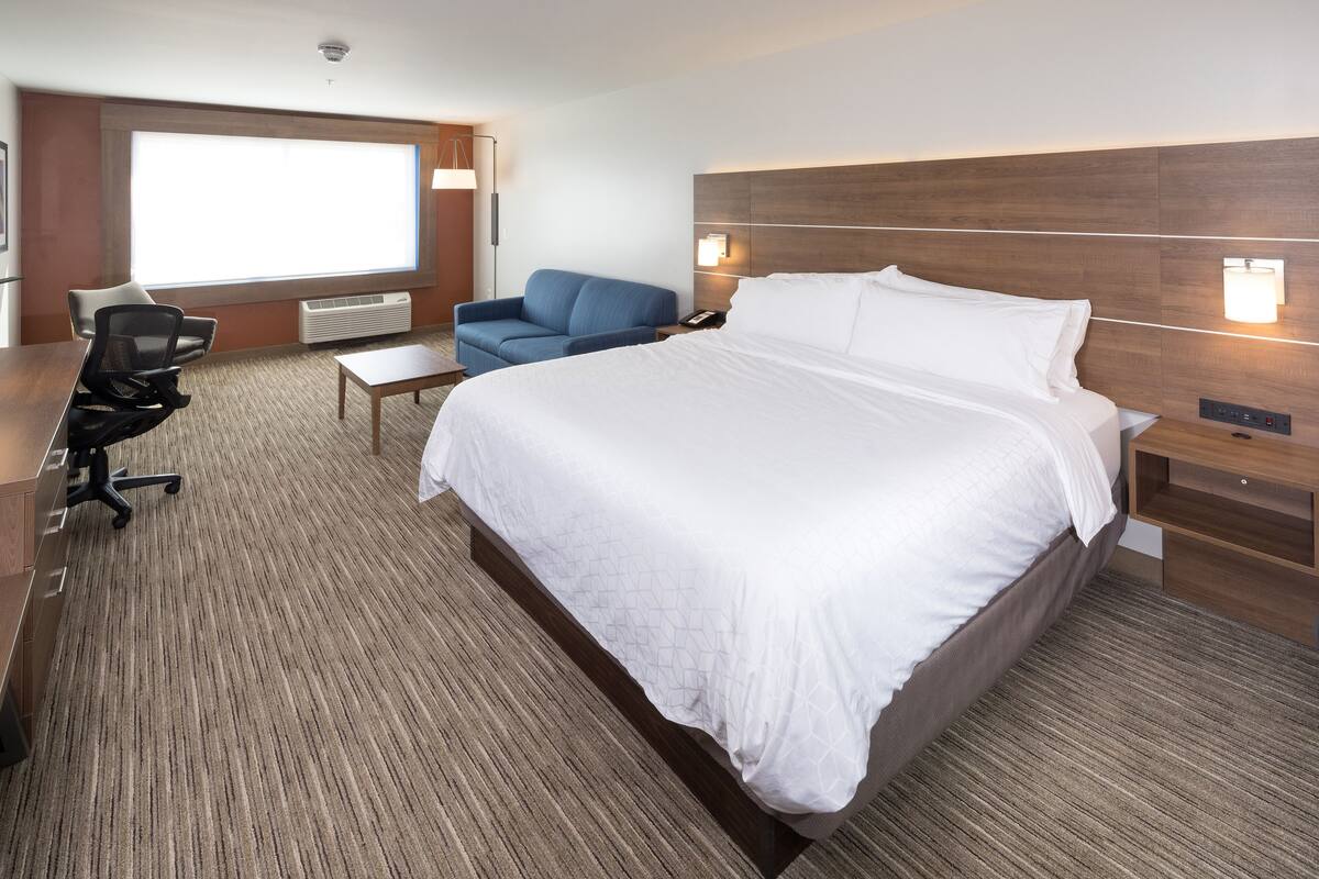 A hotel room with a large bed, a desk with a chair, a blue sofa, and a large window providing natural light.