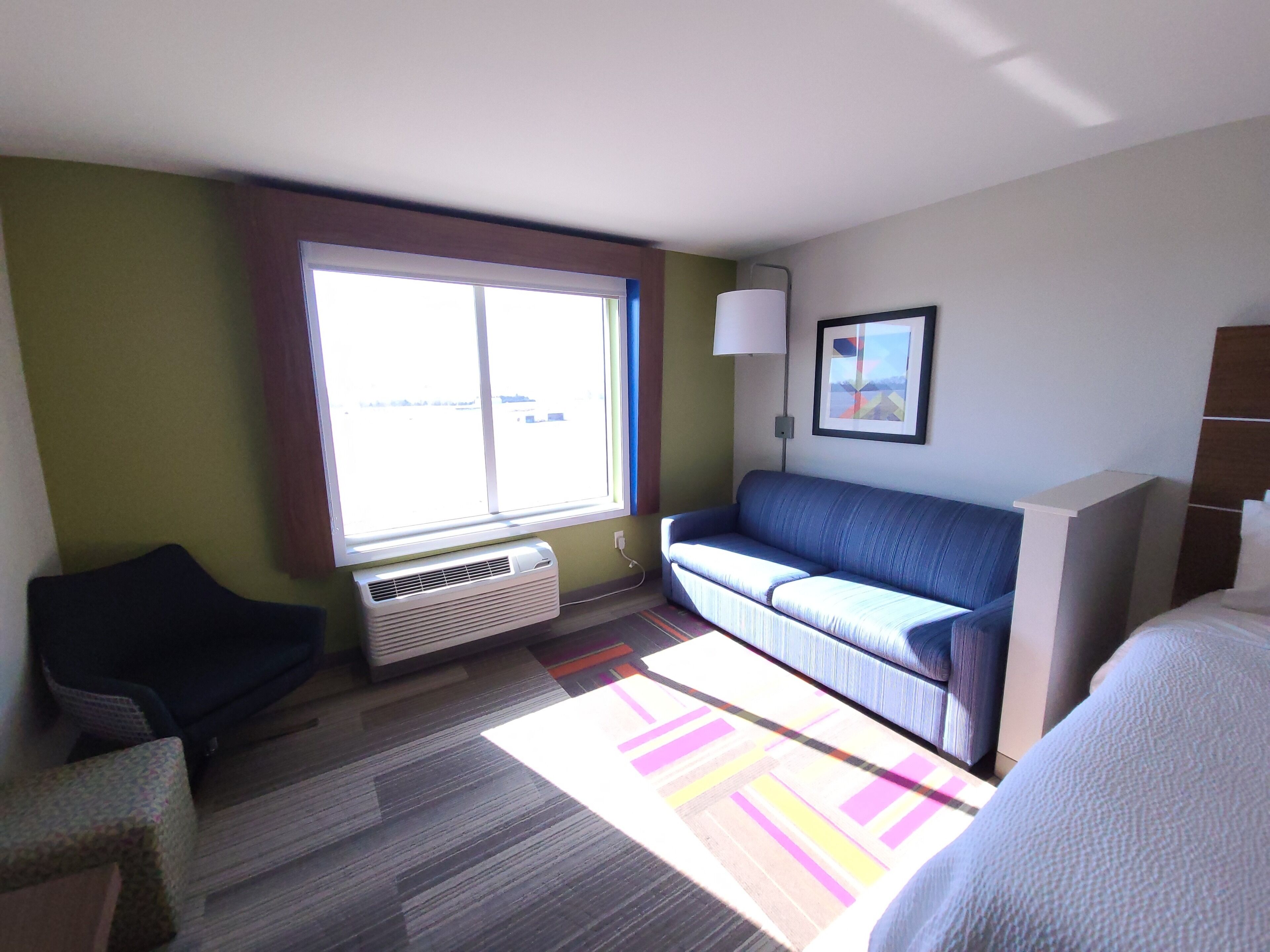 Habitación de hotel con sofá, sillón, cama, ventana con persianas, lámpara de pared y un cuadro enmarcado.