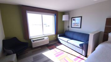 Habitación de hotel con sofá, sillón, cama, ventana con persianas, lámpara de pared y un cuadro enmarcado.