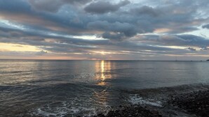 Een strand met kalm water dat de lucht weerspiegelt, een rotsachtige kustlijn en verre boten.