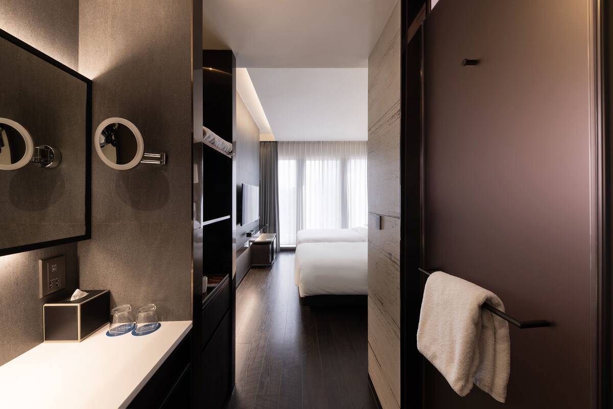 A modern hotel room with a large bed, a bedside table with a lamp, a mirror, and a towel hanging on the door.