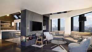 Una sala moderna con un sofá seccional, una mesa de centro y un televisor montado en la pared. Se aprecia una vista panorámica de la ciudad a través de amplias ventanas.