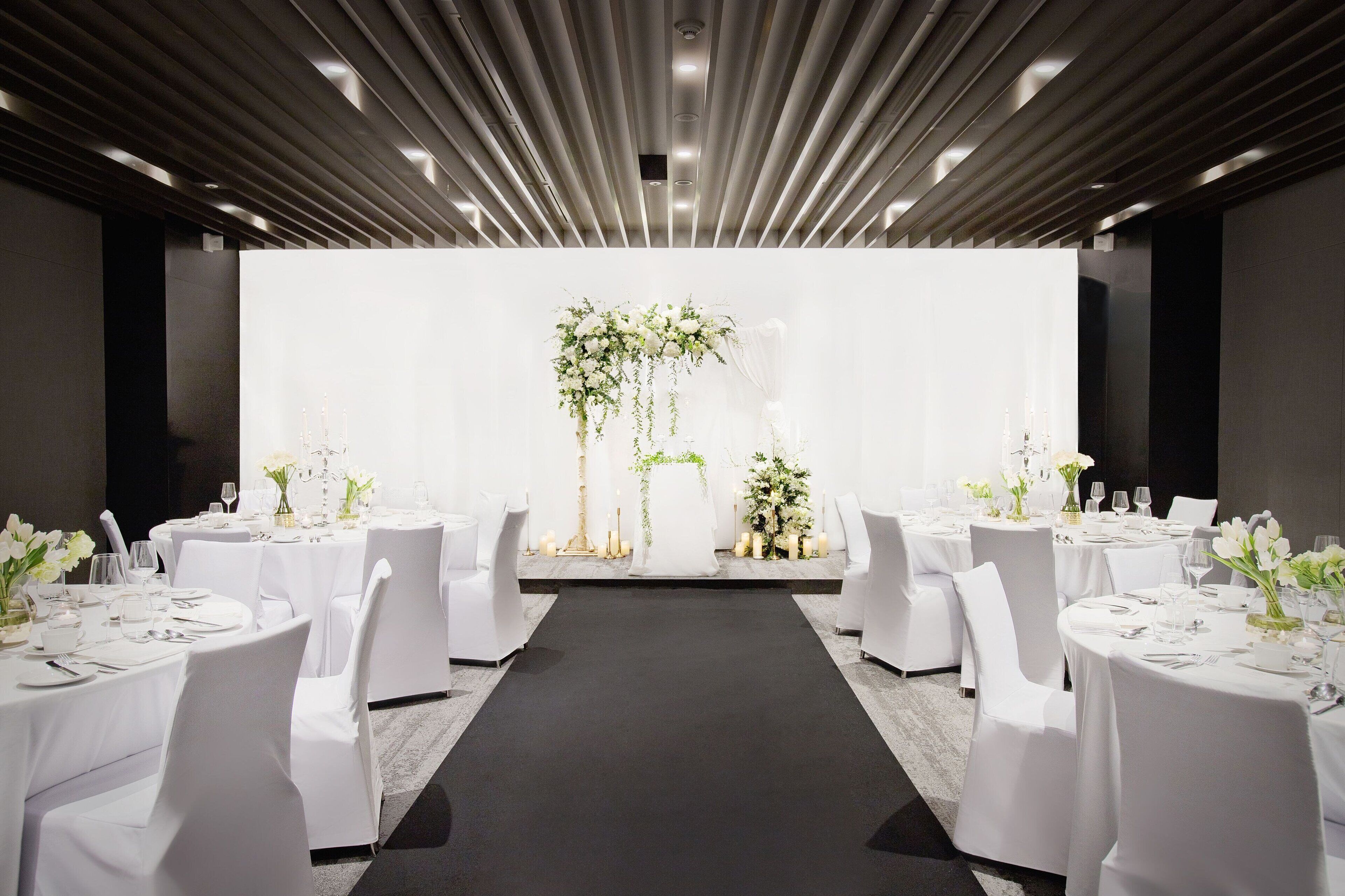 Sala de conferências com mesas redondas, cadeiras brancas e um corredor central que leva a um palco com decoração floral.
