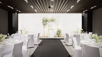 Sala de conferências com mesas redondas, cadeiras brancas e um corredor central que leva a um palco com decoração floral.