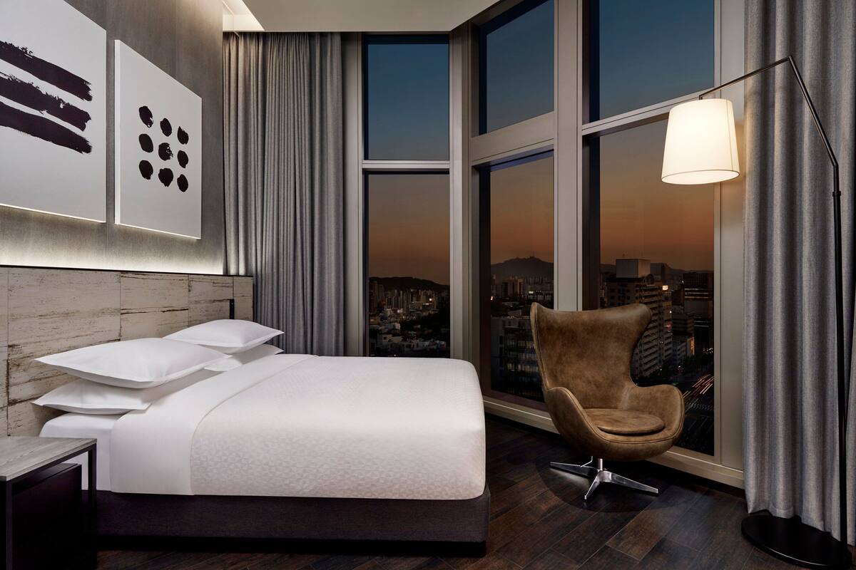 A modern hotel room with a large bed, a city view, and a contemporary chair.