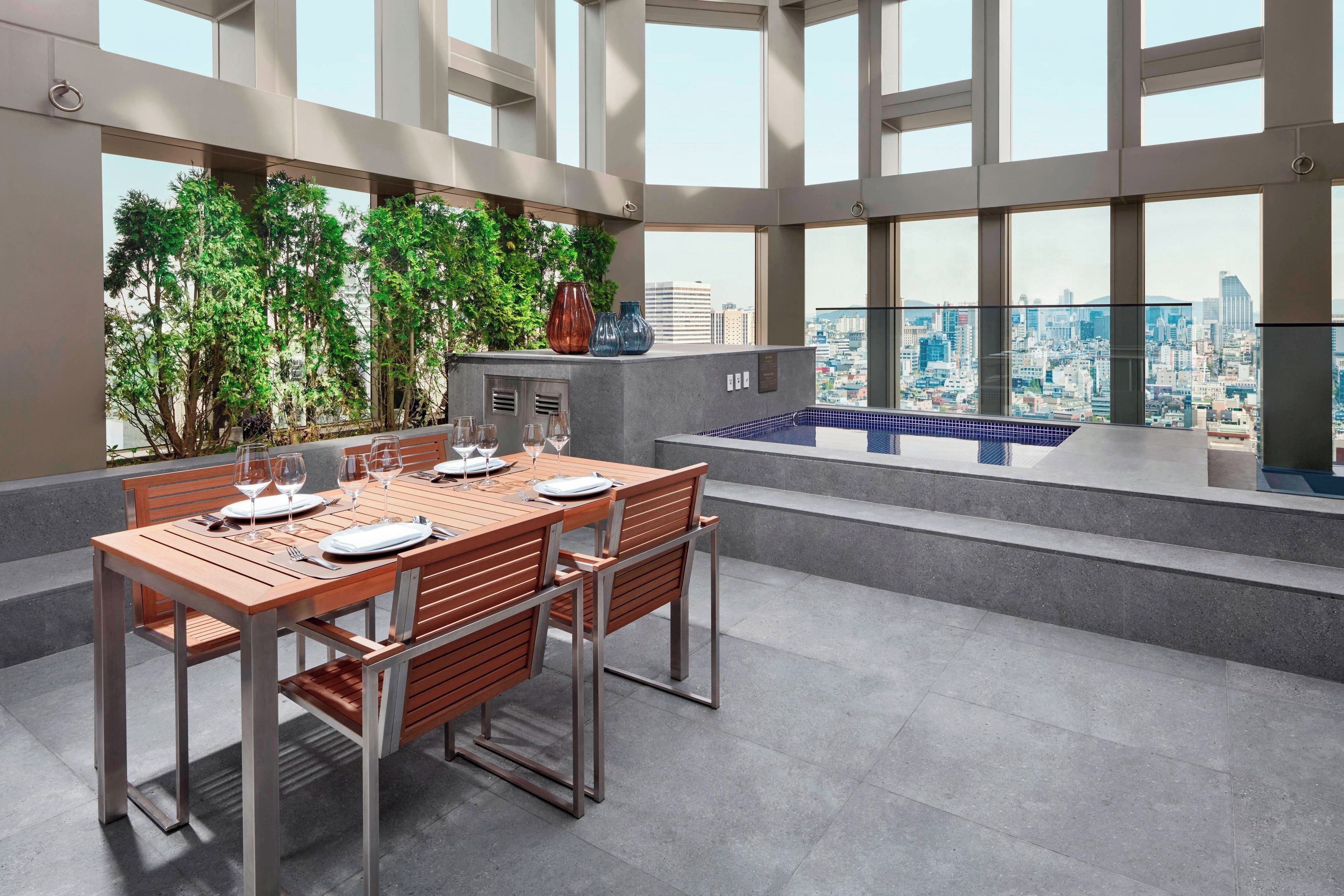 Un comedor con una mesa dispuesta para cuatro personas, vistas a la ciudad a través de amplias ventanas y una piscina pequeña interior.
