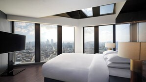 Habitación de hotel moderna con una cama grande, televisión y vistas a la ciudad a través de amplios ventanales.