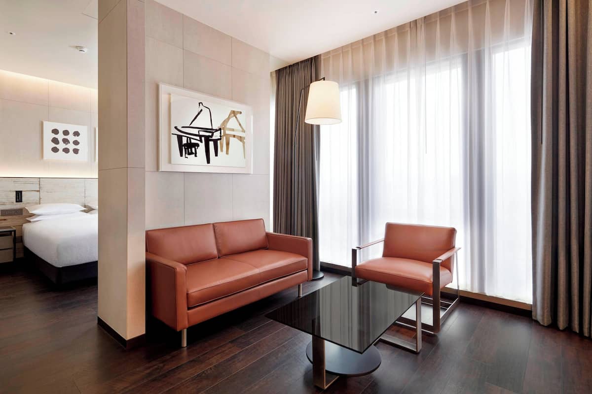 A modern hotel room with a large bed, a sofa, a chair, a glass coffee table, and a large window with curtains.