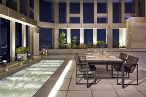 Terraza en la azotea con jacuzzi, mesa de comedor y sillas, con vista a un paisaje urbano nocturno.