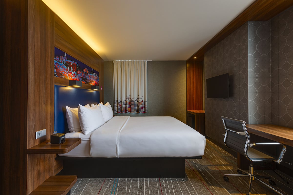 Una habitación de hotel moderna con una cama grande, un escritorio, una silla, un televisor de pantalla plana y un elemento decorativo en la pared.