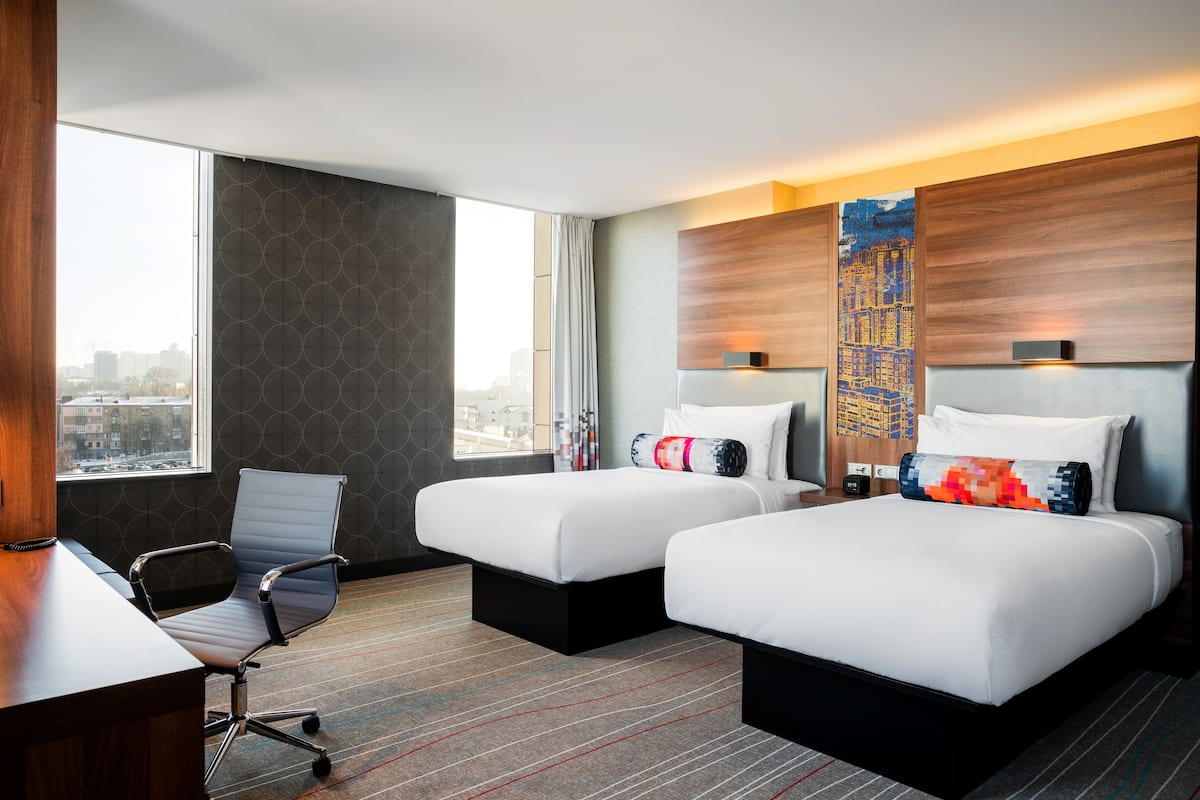 Habitación de hotel con dos camas, un escritorio, una silla y un ventanal que ofrece vistas a la ciudad.