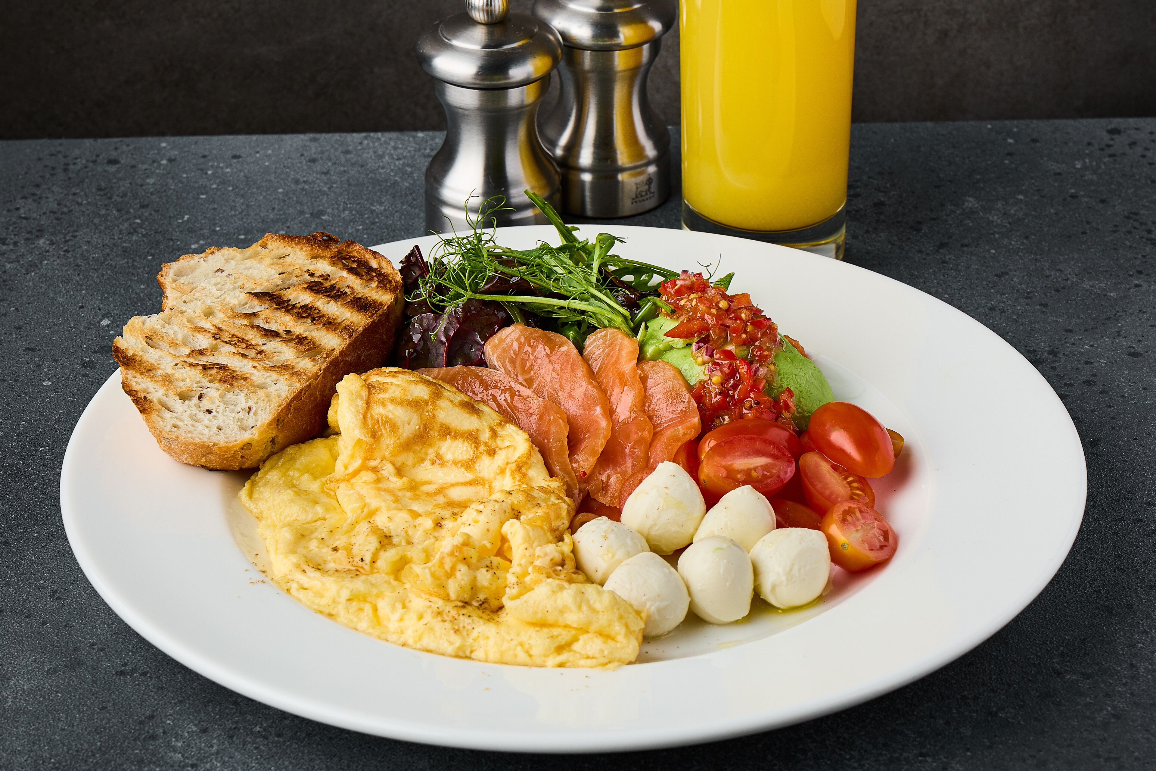Un plato de comida para el desayuno con tostadas, huevos revueltos, salmón ahumado, tomates, bolitas de mozzarella y vegetales verdes.
