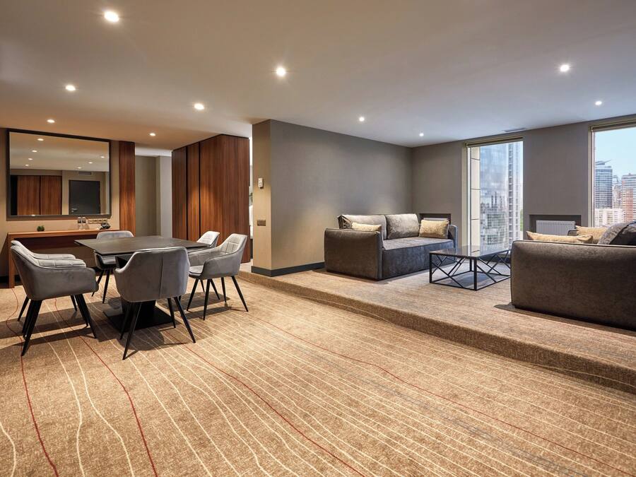 A modern hotel room with a dining area, a sofa set, and a large window offering a city view.