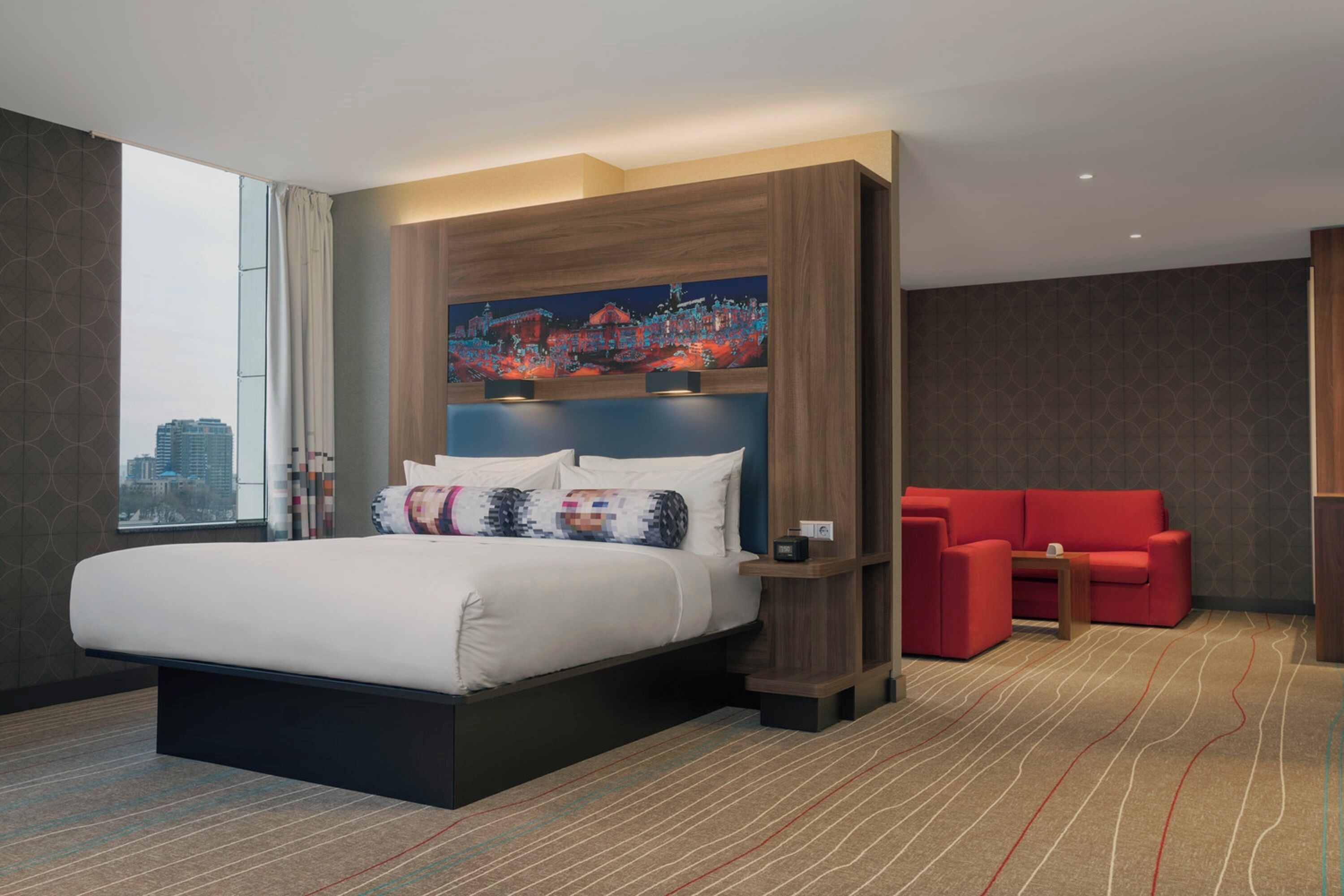 Ein modernes Hotelzimmer mit einem großen Bett, einem roten Sofa, einem Stadtbild an der Wand und Blick auf die Stadt durch das Fenster.