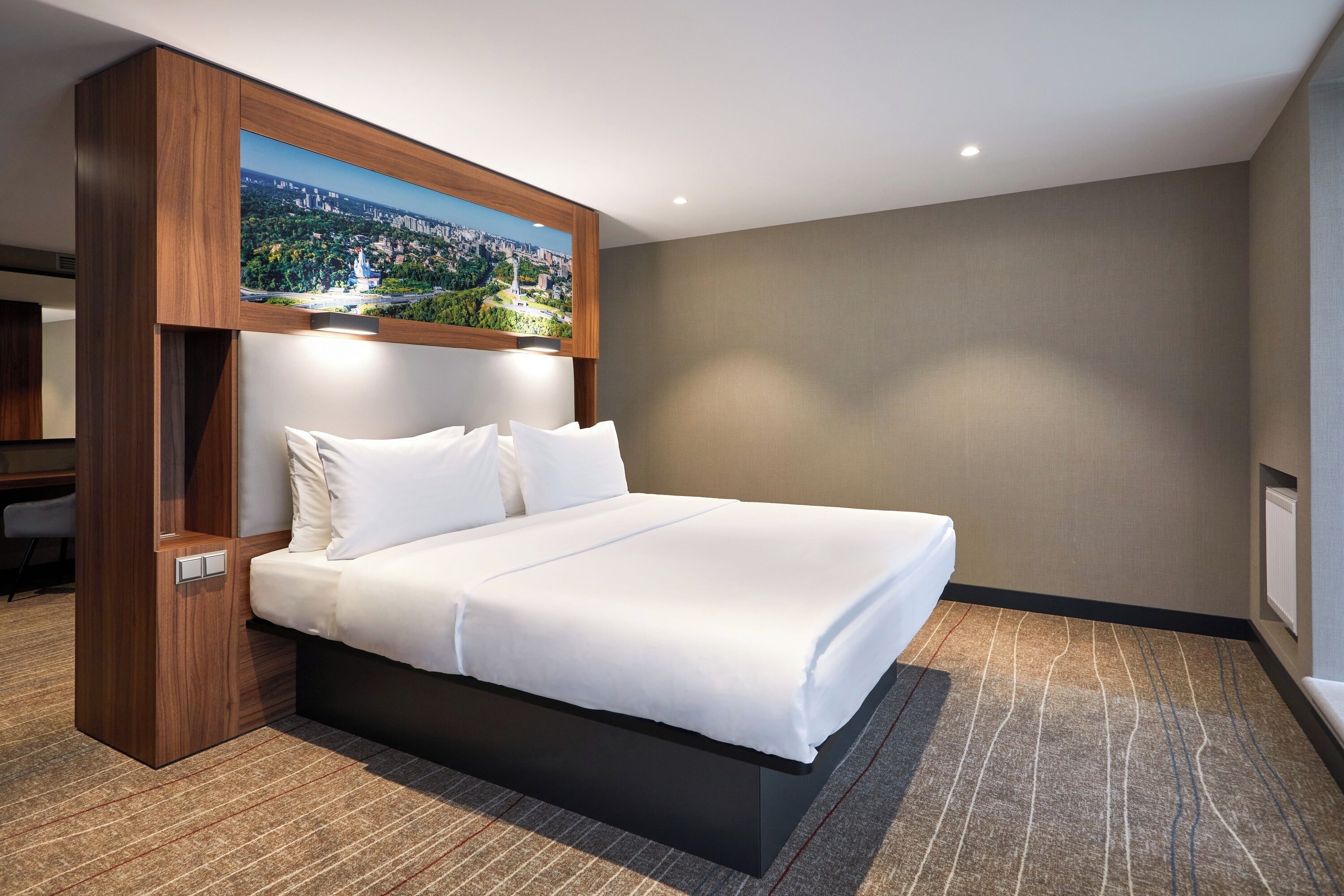 A hotel room with a large bed, a wooden headboard, a cityscape painting above the bed, and a carpeted floor.
