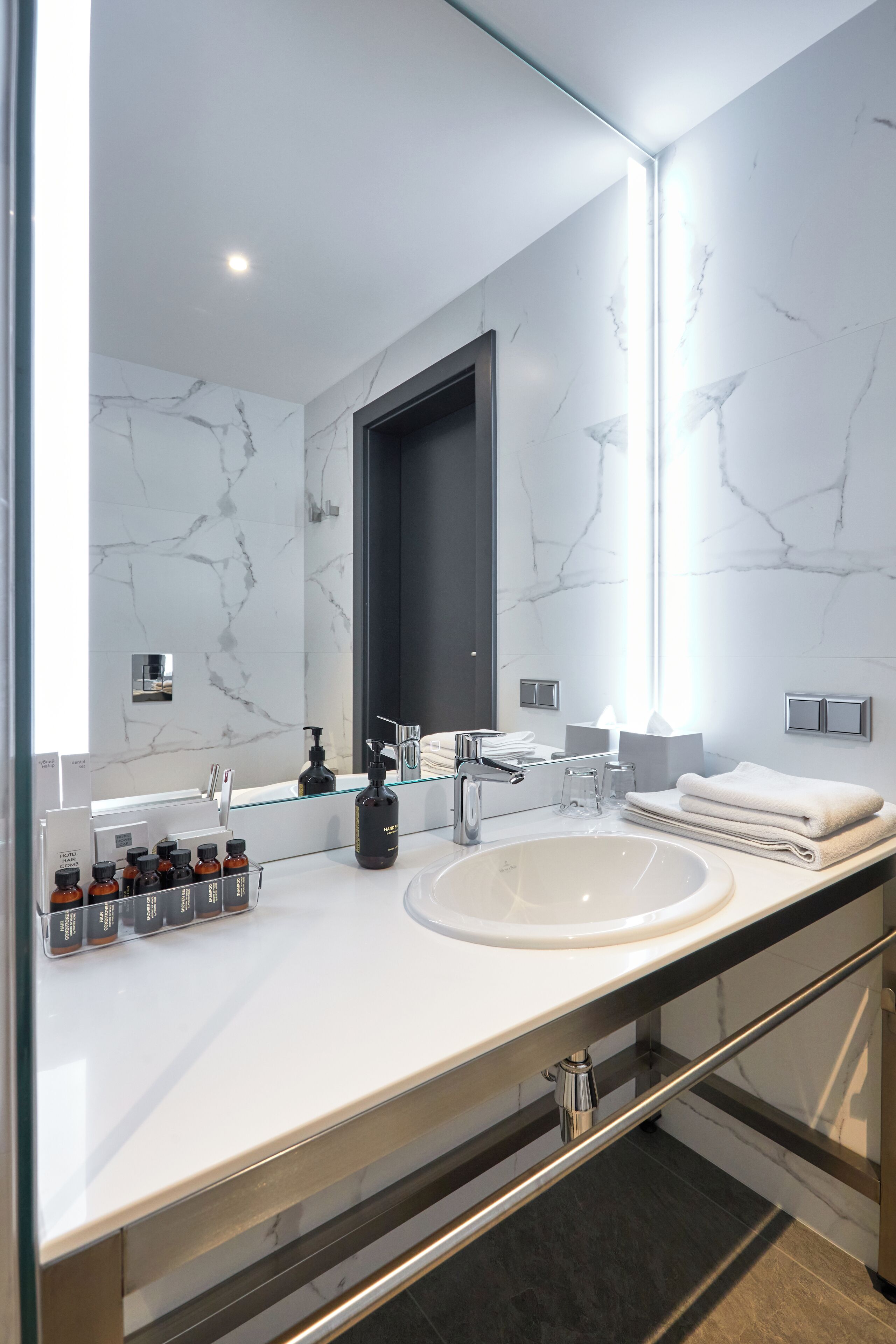 Un baño moderno con un espejo grande, paredes de mármol blanco, un lavamanos blanco y una cabina de ducha con paredes de vidrio.